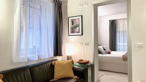 Ein gemütliches Wohnzimmer mit einem Ledersofa, einem Tisch mit einer Schale, einem gerahmten Bild an der Wand und einem Durchgang zu einem Schlafzimmer mit Bett und Vorhängen.