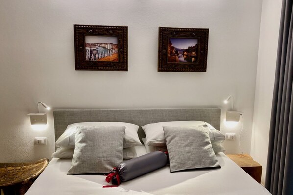 A neatly made bed with two pillows and a rolled blanket, a bedside table with a lamp, and two framed pictures on the wall.