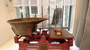 Ein alter roter Metal-Eimer auf einem Tisch, neben einem Glas bernsteinfarbener Flüssigkeit, platziert vor einem Fenster mit Blick auf ein Ziegelgebäude.