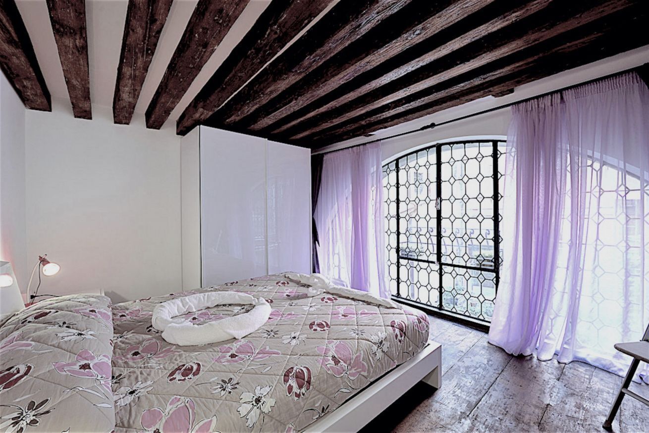 Una camera da letto con soffitto in legno, un letto con lenzuola a motivi floreali, una finestra con tende viola e vista attraverso una griglia esagonale.