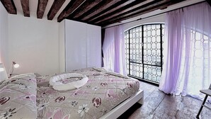 Een slaapkamer met een houten plafond, een bed met bloemenbeddengoed, een raam met paarse gordijnen en uitzicht door een zeshoekig raamrooster.