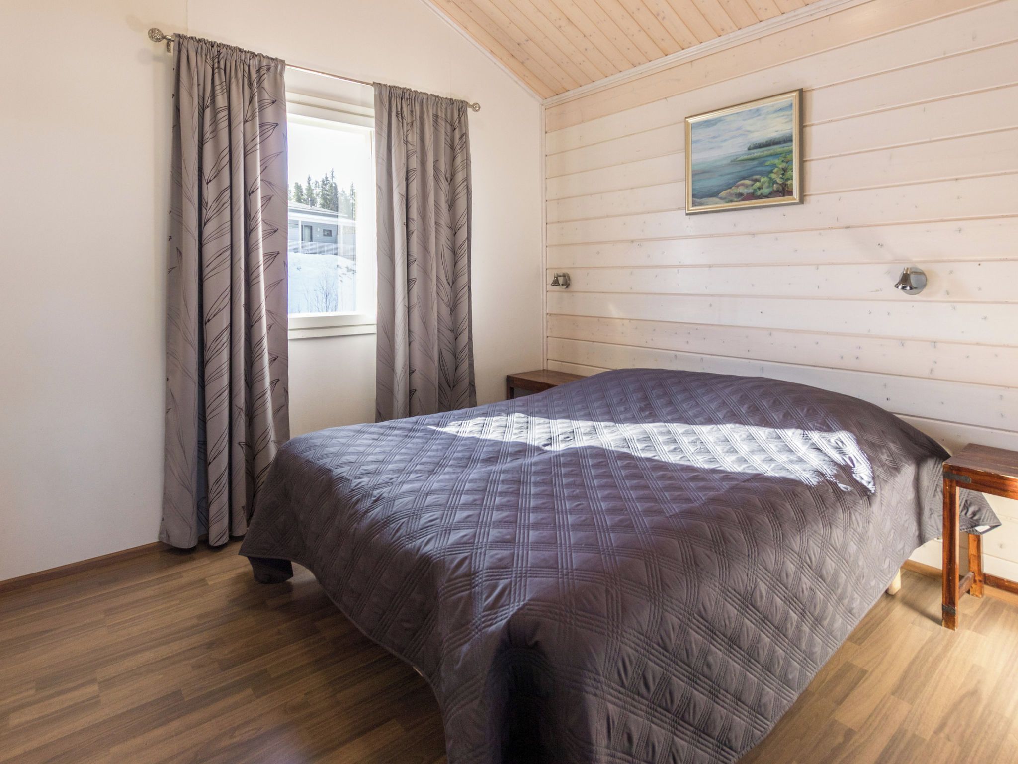 Une chambre à coucher avec un grand lit, un plancher en bois, une fenêtre avec des rideaux, un tableau au mur et une petite table de chevet.