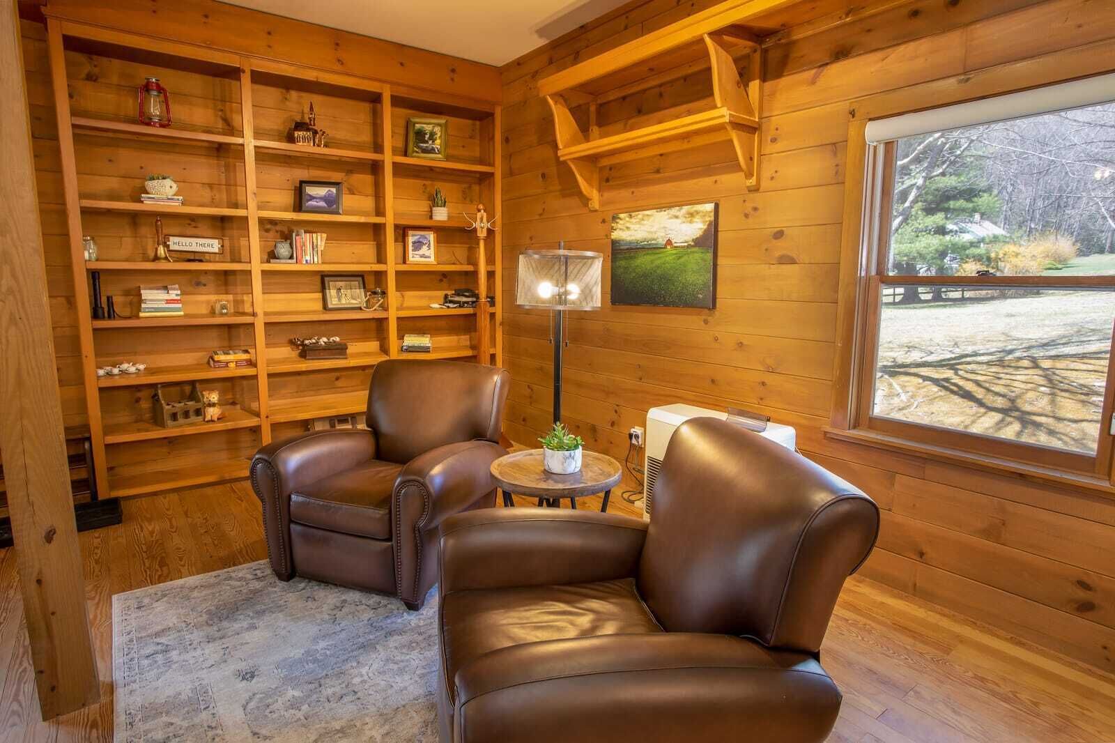 Una habitación acogedora con paredes de madera, una estantería, dos sillones de cuero, una mesita con una planta y una ventana con vistas a árboles.