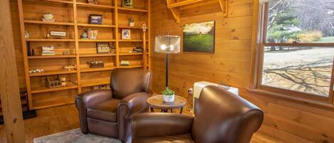 Una habitación acogedora con paredes de madera, una estantería, dos sillones de cuero, una mesita con una planta y una ventana con vistas a árboles.