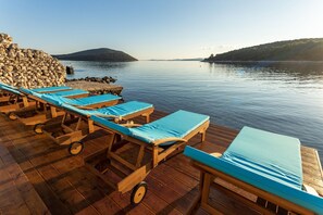 A wooden deck with lounge chairs and a stone wall overlooking calm water with distant hills. - Villa Otus ZadarVillas (Pašman)