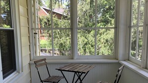 Ein überdachter Balkon mit Holztisch und zwei Stühlen, große Fenster mit weißem Zierrahmen und einem hängenden Dekorationsobjekt.