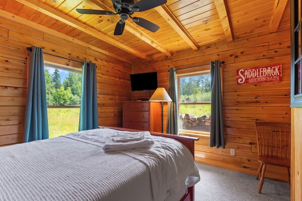 A wooden cabin room with a bed, a chair, a TV, and a sign reading "Saddleback Mountain".
