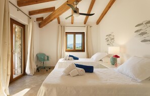 Ein Hotelzimmer mit zwei Betten, einem Deckenventilator, einem Fenster mit Blick auf die Berge, einem Schreibtisch mit Lampe und einem Sessel.