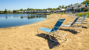 A beach with rows of blue and white sun loungers facing a calm lake, surrounded by a row of houses with a clear view of the water.