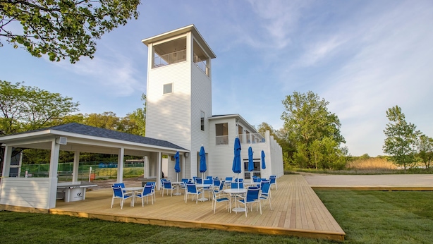 A white building with a tower, a covered patio with blue chairs and umbrellas, and a grassy deck.