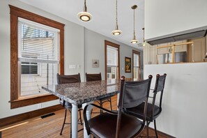 A modern kitchen with a marble countertop, wooden chairs, pendant lighting, and a view through the window.