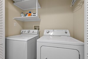 A laundry room with two white front-loading washers and dryers, a shelf with cleaning supplies, and a white door.