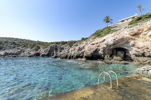 A rocky coastline with clear blue water, a stone staircase leading to a building, and a palm tree. - Beautiful 3-bed Sea Front Villa in Agios Nikolaos (Agios Nikolaos)