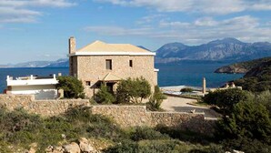 A stone house with a tiled roof, situated on a hillside with a view of the sea and mountains in the background. - Beautiful 3-bed Sea Front Villa in Agios Nikolaos (Agios Nikolaos)