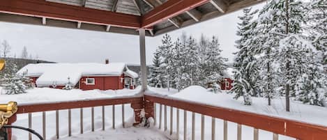 Ein überdachter Außenbereich mit Holzterrasse, Metallgeländer und Blick auf schneebedeckte Bäume sowie ein rotes Gebäude im Hintergrund.