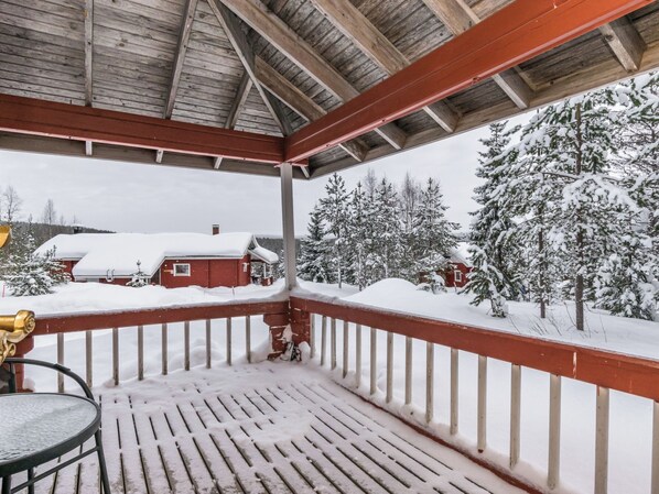Un espacio al aire libre techado con una terraza de madera, baranda metálica y vista a árboles cubiertos de nieve y un edificio rojo al fondo.