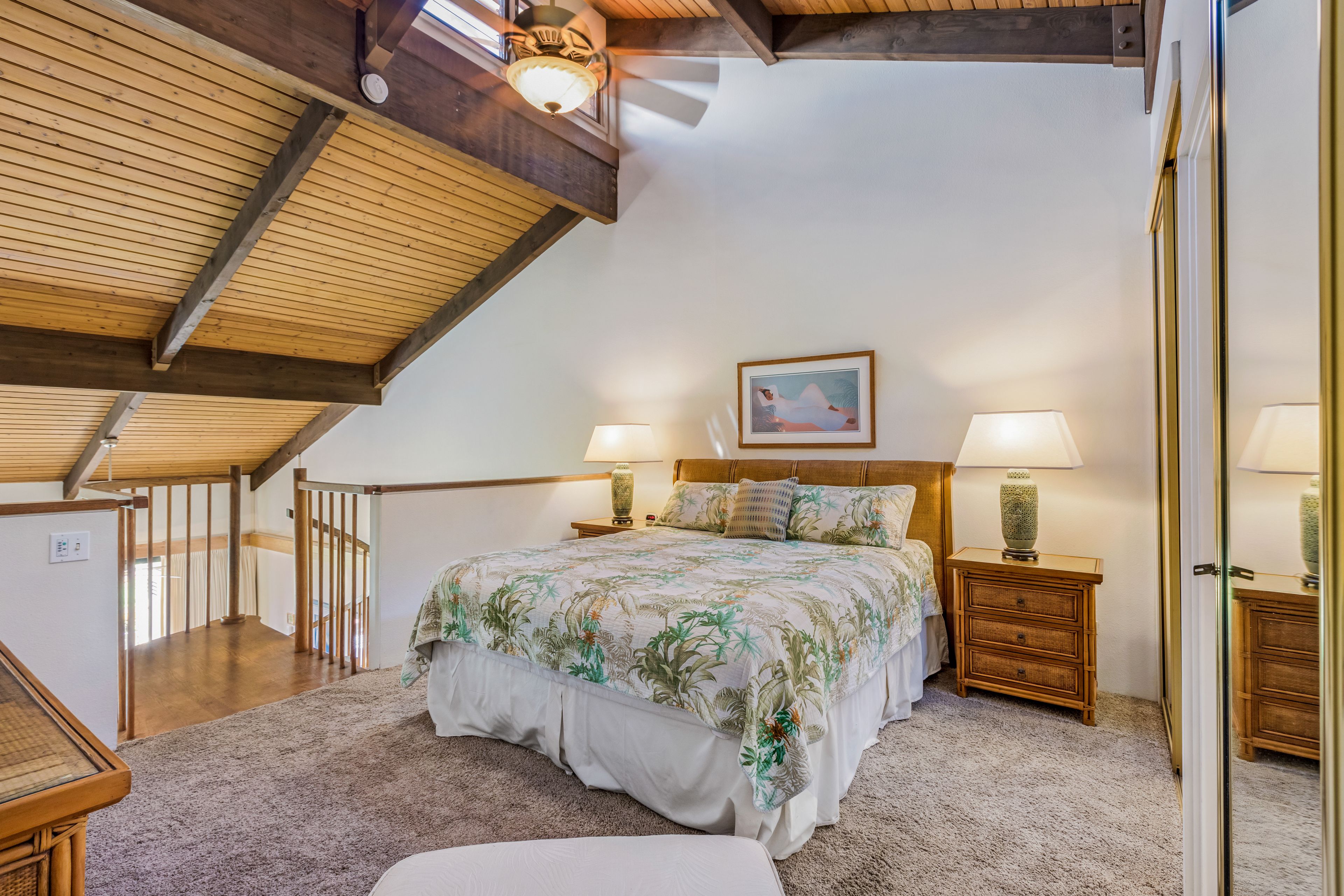 Ein Schlafzimmer mit schrägen Wänden, Holzbalken, einem großen Bett mit Blumenmuster auf der Bettwäsche, zwei Nachttischen mit Lampen und einem gerahmten Bild an der Wand.