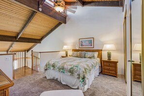A bedroom with a sloped ceiling, wooden beams, a large bed with floral bedding, two bedside tables with lamps, and a framed picture on the wall.