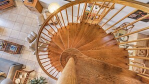 Una escalera en espiral de madera en una habitación luminosa con piso de baldosas, rodeada de muebles y objetos decorativos.