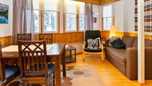 A cozy living room with wooden walls, a dining table, chairs, a sofa, and a large window with curtains.