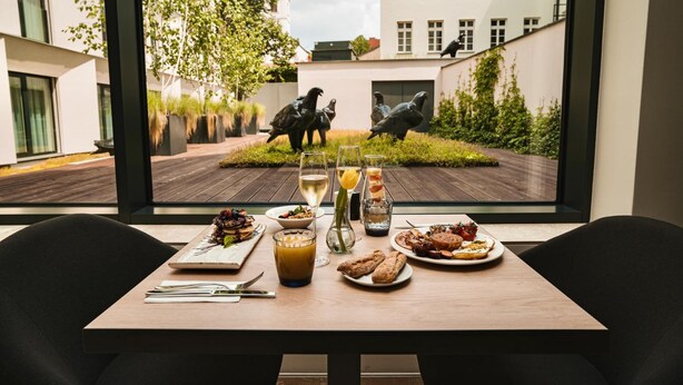 Ein Esstisch mit Mahlzeit und Getränken, Blick auf einen Innenhof mit Statuen und moderne Gebäude im Hintergrund.
