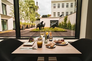 A dining table with a meal and drinks, a view of a courtyard with statues, and modern buildings in the background.