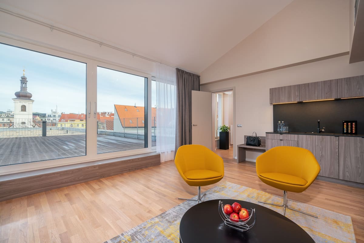 A modern living room with large windows, two yellow chairs, a black table with a fruit bowl, and a kitchen area with wooden cabinets.