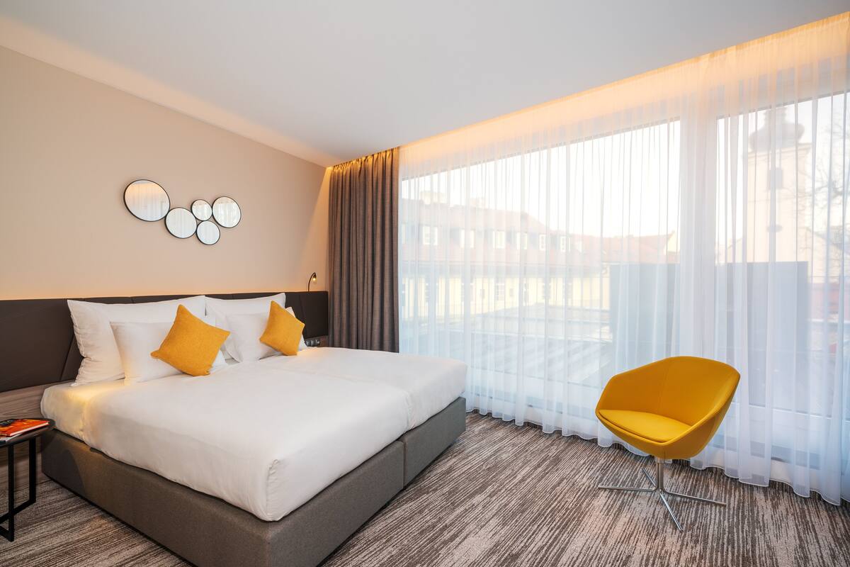 A hotel room with a large bed, a modern chair, and a view through sheer curtains.