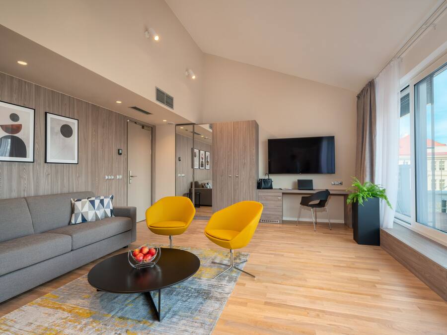 Ein modernes Wohnzimmer mit einer Couch, zwei gelben Sesseln, einem Couchtisch mit Früchten, einem Fernseher und einem großen Fenster mit Aussicht.