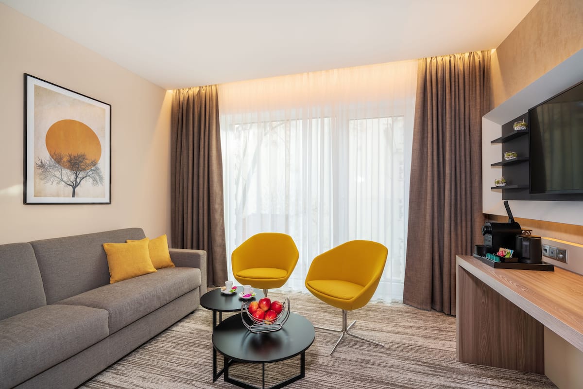 A modern hotel room with a grey sofa, two yellow armchairs, a coffee table with fruit, a flat-screen TV, and a large window with curtains.