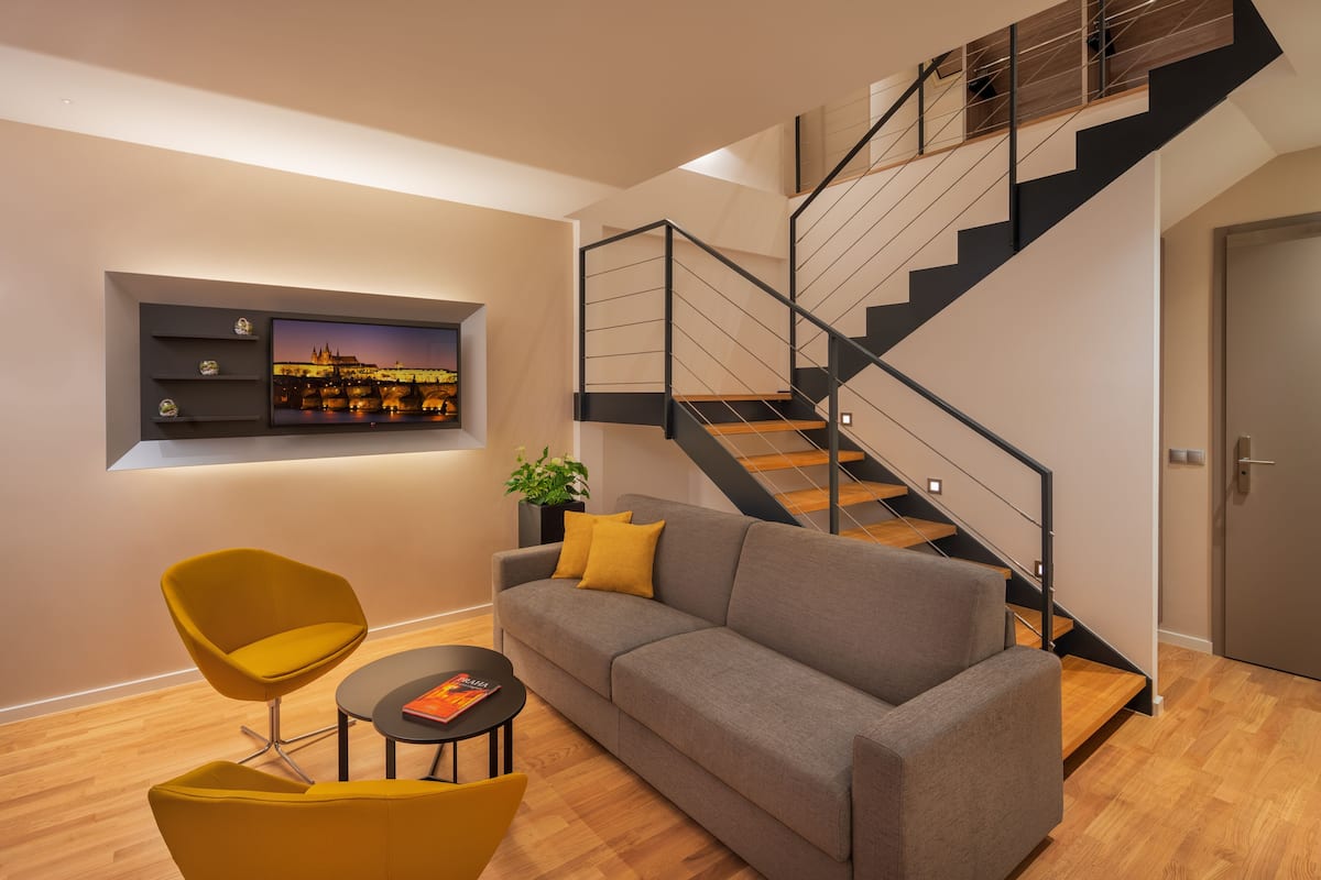 A modern living room with a grey sofa, yellow chairs, a flat-screen TV, and a staircase with wooden steps and black railings.