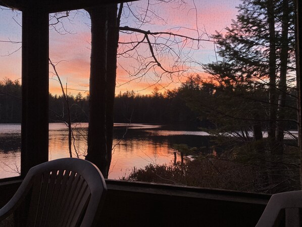 A view from a covered porch overlooking a lake with trees and a sunset.