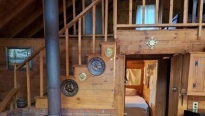 Interno di una rustica baita con stufa a legna, camera da letto soppalcata e una scala adornata con piatti decorativi.