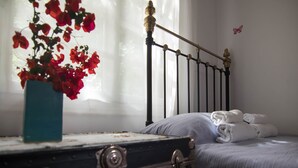 A bed with white linens, a blue vase with red flowers, and a window with sheer curtains. - Kitries Home (West Mani)