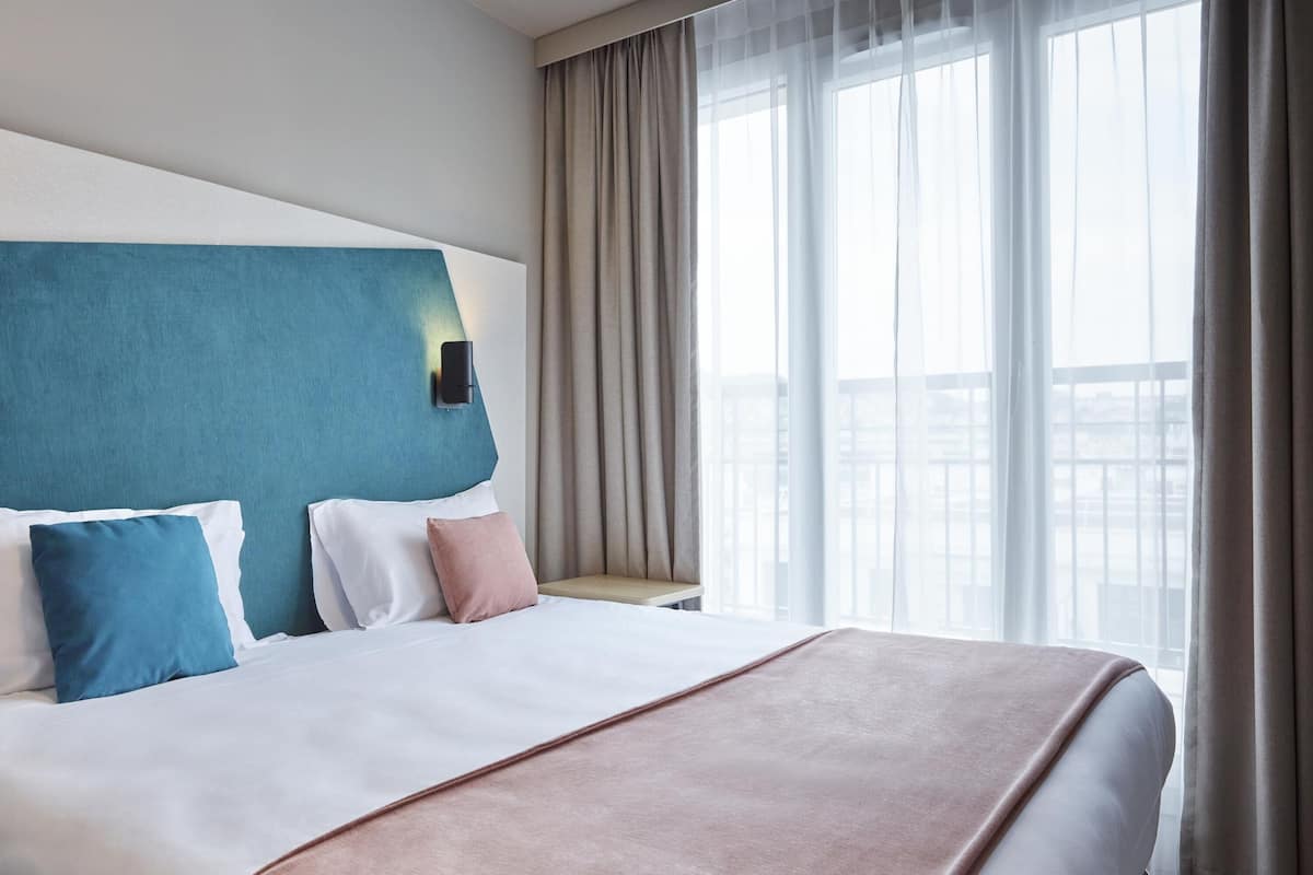 Une chambre d’hôtel avec un grand lit, une tête de lit, une table de chevet, une fenêtre avec des rideaux et une vue sur la ville.