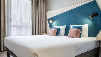 Une chambre d’hôtel avec un grand lit, une tête de lit agrémentée d’une touche de bleu et une fenêtre avec des rideaux.
