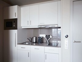 Cocina compacta con armarios blancos, microondas integrado, electrodomésticos de acero inoxidable y una encimera con hervidor y utensilios.