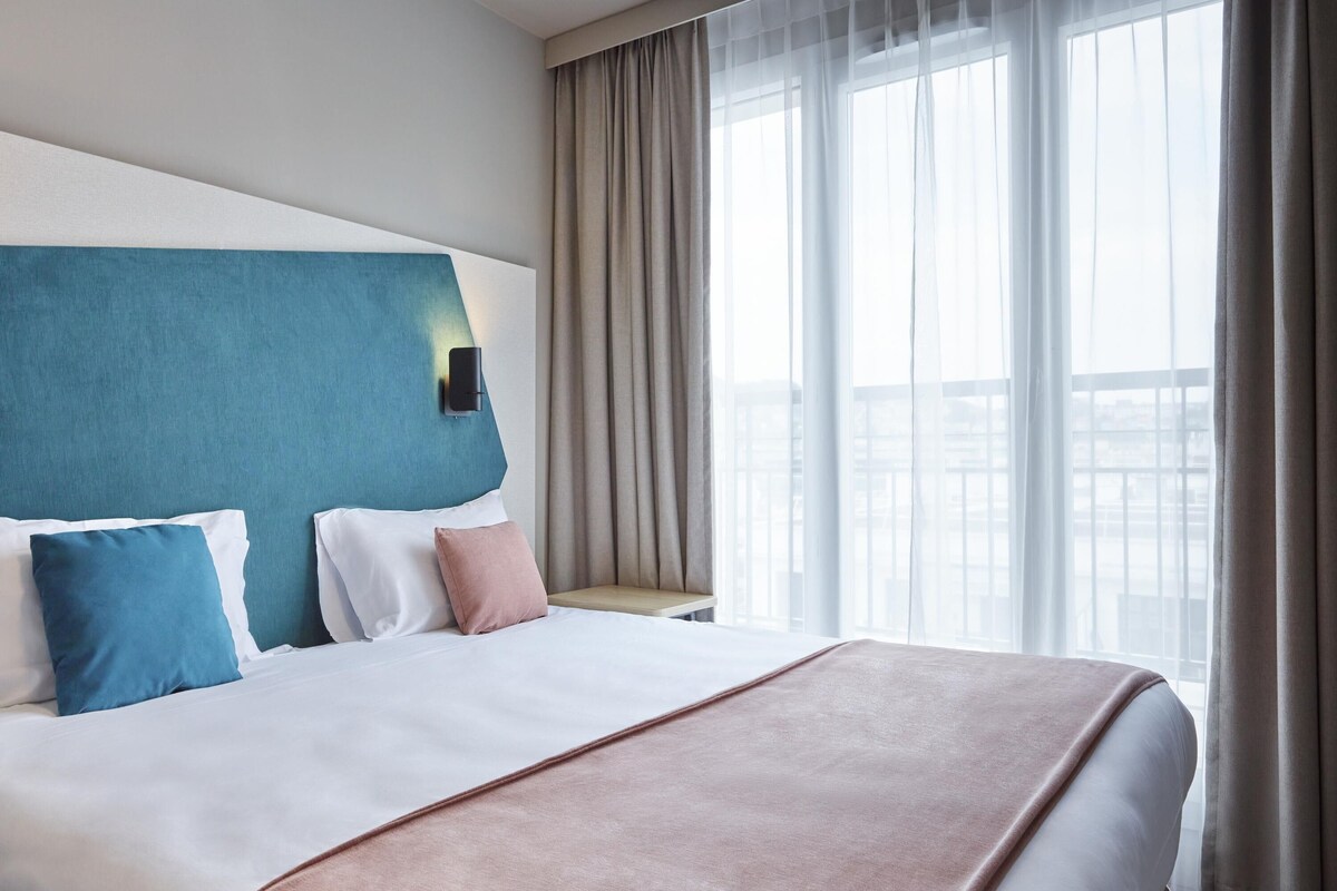 Habitación de hotel con una cama grande, cabecera, mesita de noche, ventana con cortinas y vistas a la ciudad.