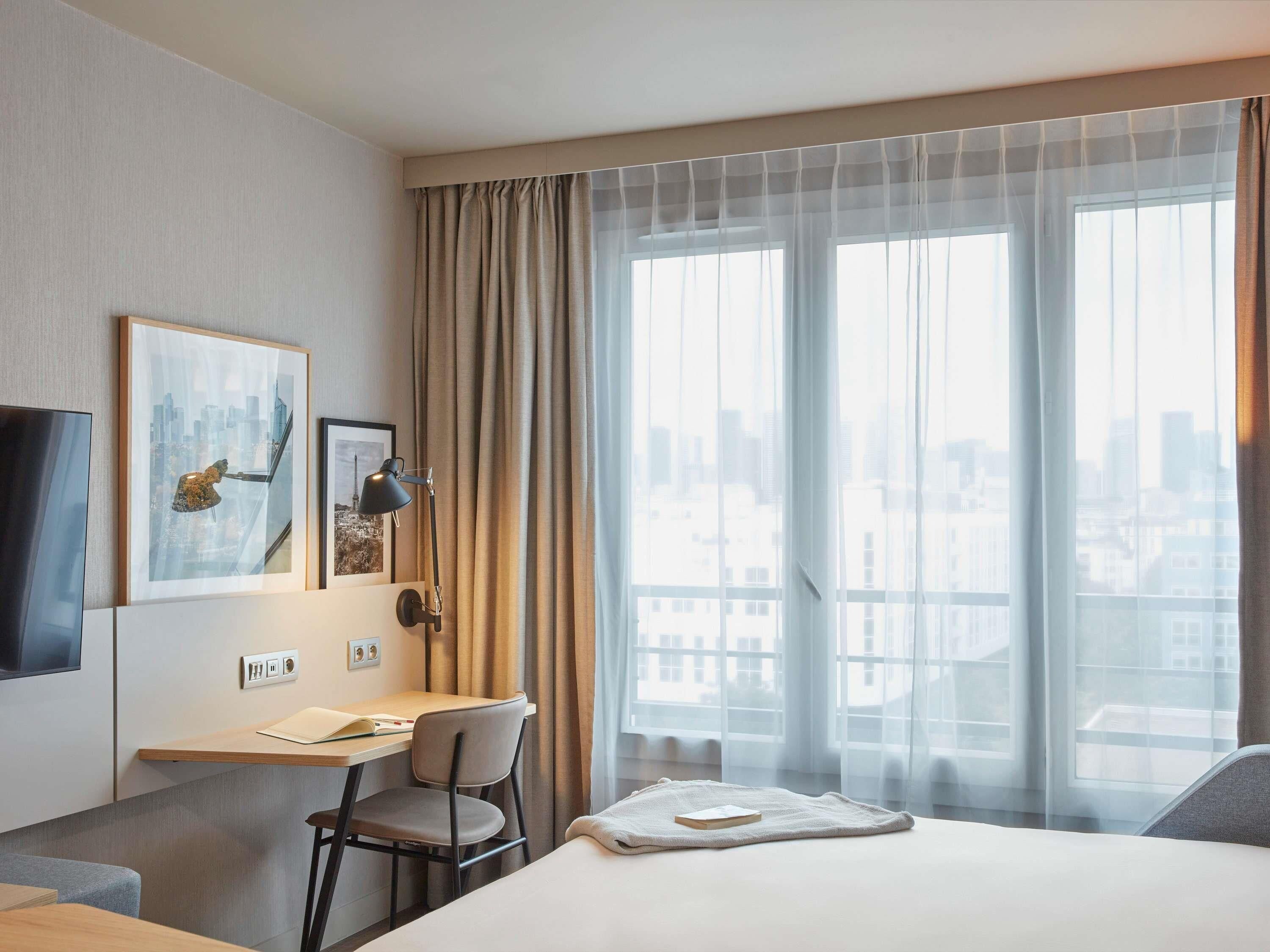 A hotel room with a bed, a desk with a chair, a TV, and a large window with curtains.