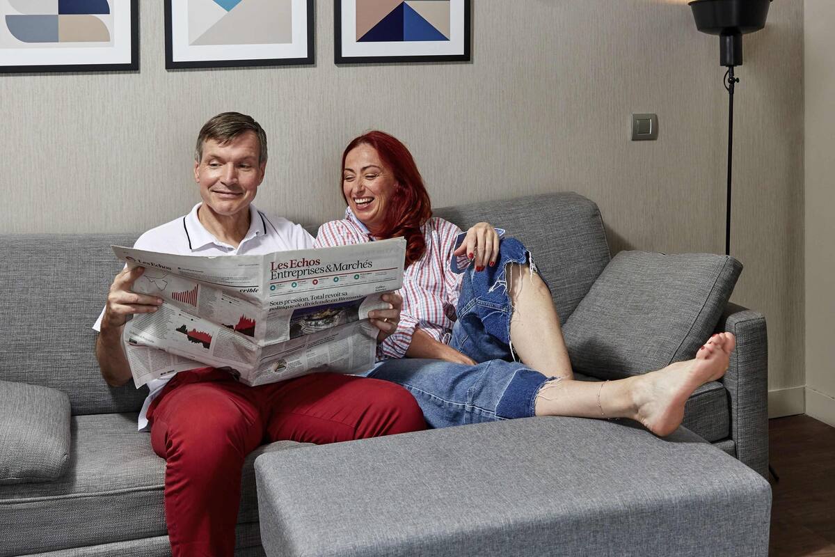 Un couple est assis sur un canapé gris, avec un journal, une lampe et une œuvre d’art abstrait accrochée au mur.