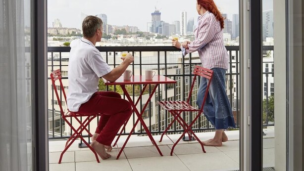 Un balcón con vista a la ciudad, una mesa con sillas y una persona sosteniendo una bebida.