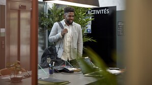 Un hombre con camisa blanca y blazer gris de pie en una oficina, con una computadora portátil, una calculadora y un bolígrafo sobre el escritorio.
