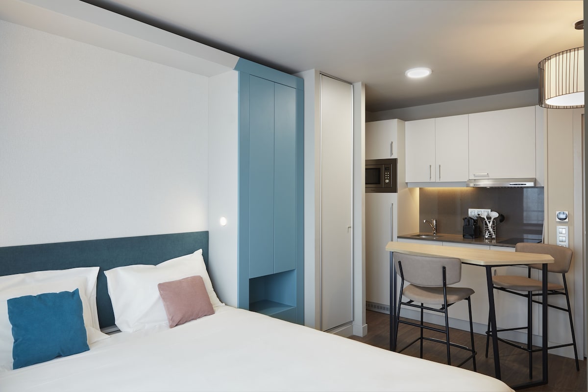 Una habitación de hotel moderna con cama, una kitchenette pequeña y una zona de comedor con dos sillas.