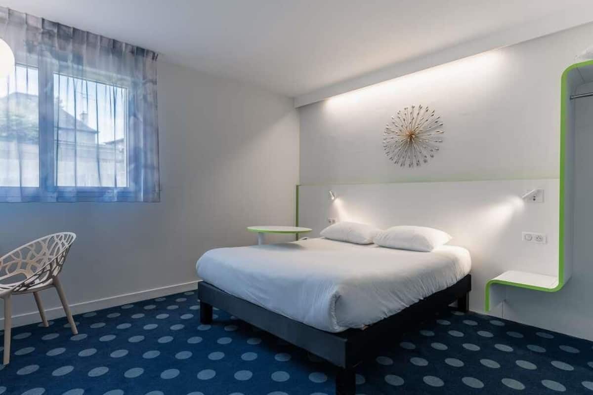 A modern hotel room with a bed, a chair, a small table, and a decorative wall piece.