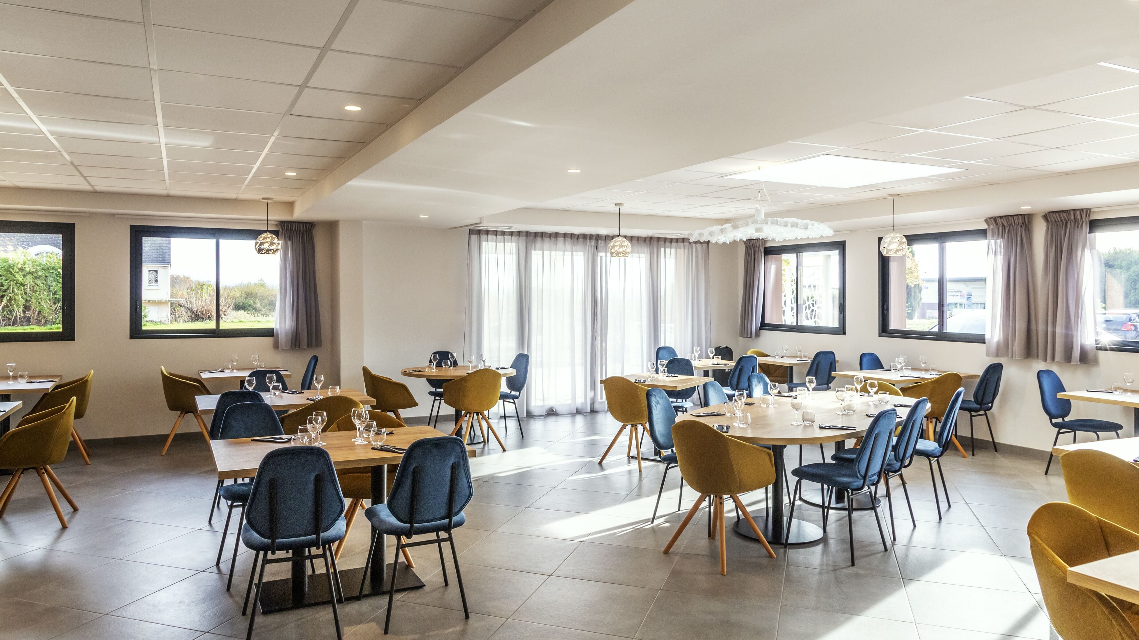 A spacious dining area with multiple tables and chairs, large windows with curtains, and a modern ceiling design.