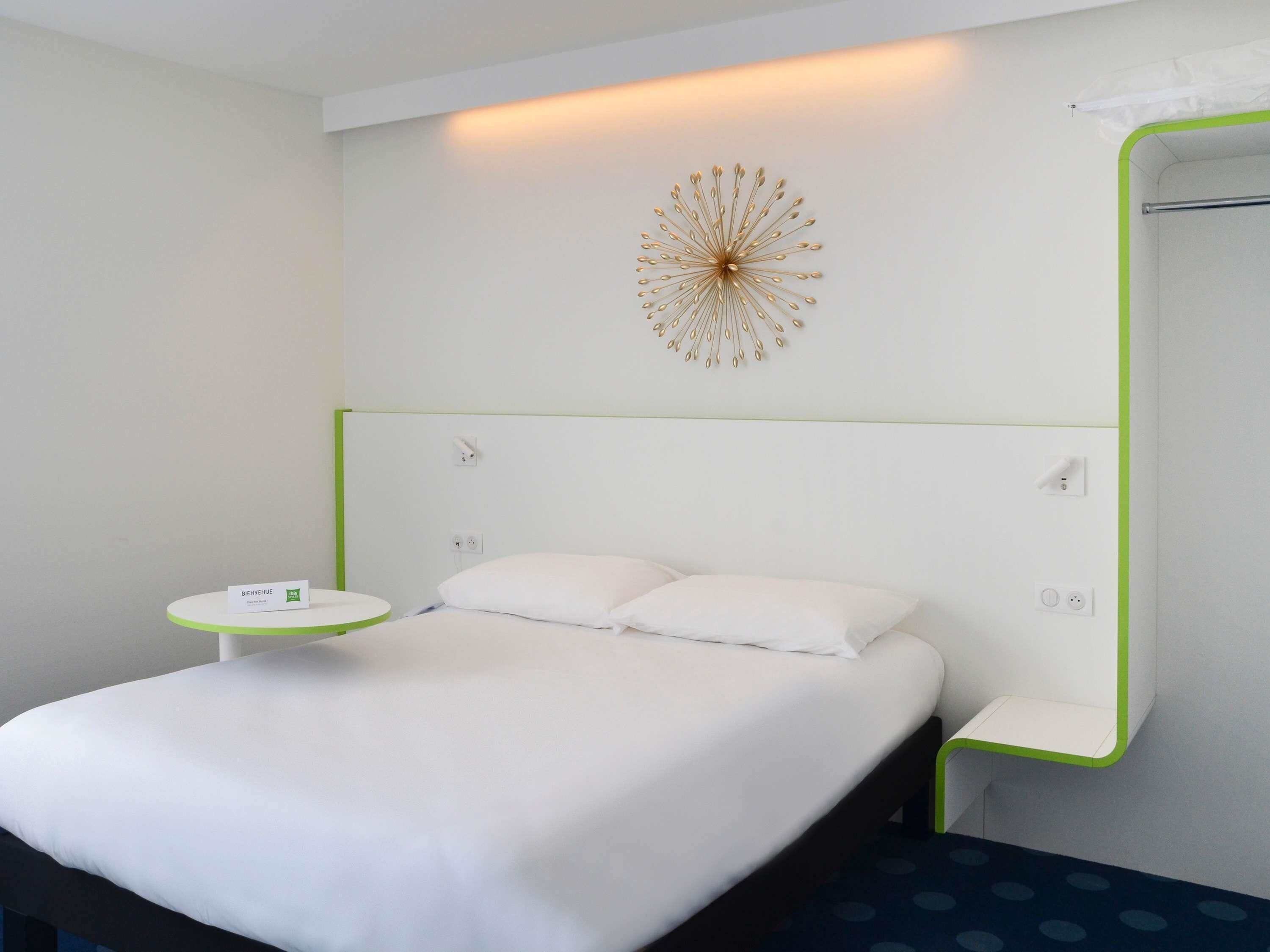 A hotel room with a bed, a bedside table, a wall-mounted decorative piece, and a mirror with a green frame.