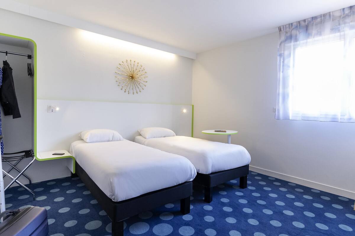 Una habitación de hotel con dos camas, una mesita, un espejo y un ventanal con cortinas.