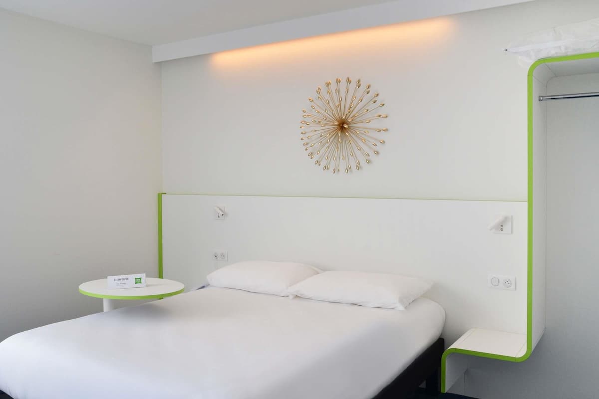 A hotel room with a bed, a bedside table, a wall-mounted decorative piece, and a mirror with a green frame.