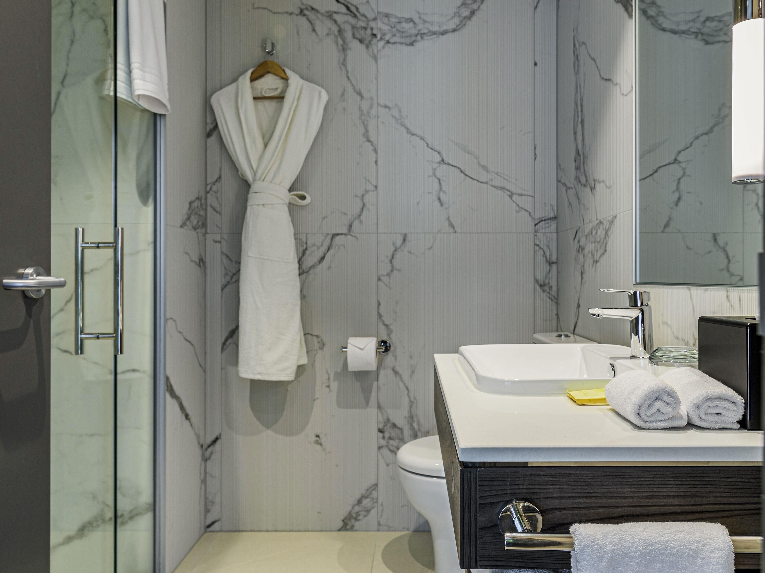 Un baño moderno con pared de mármol, cabina de ducha de vidrio, lavamanos blanco con mueble negro y una bata colgada.
