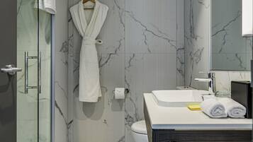 Un baño moderno con pared de mármol, cabina de ducha de vidrio, lavamanos blanco con mueble negro y una bata colgada.